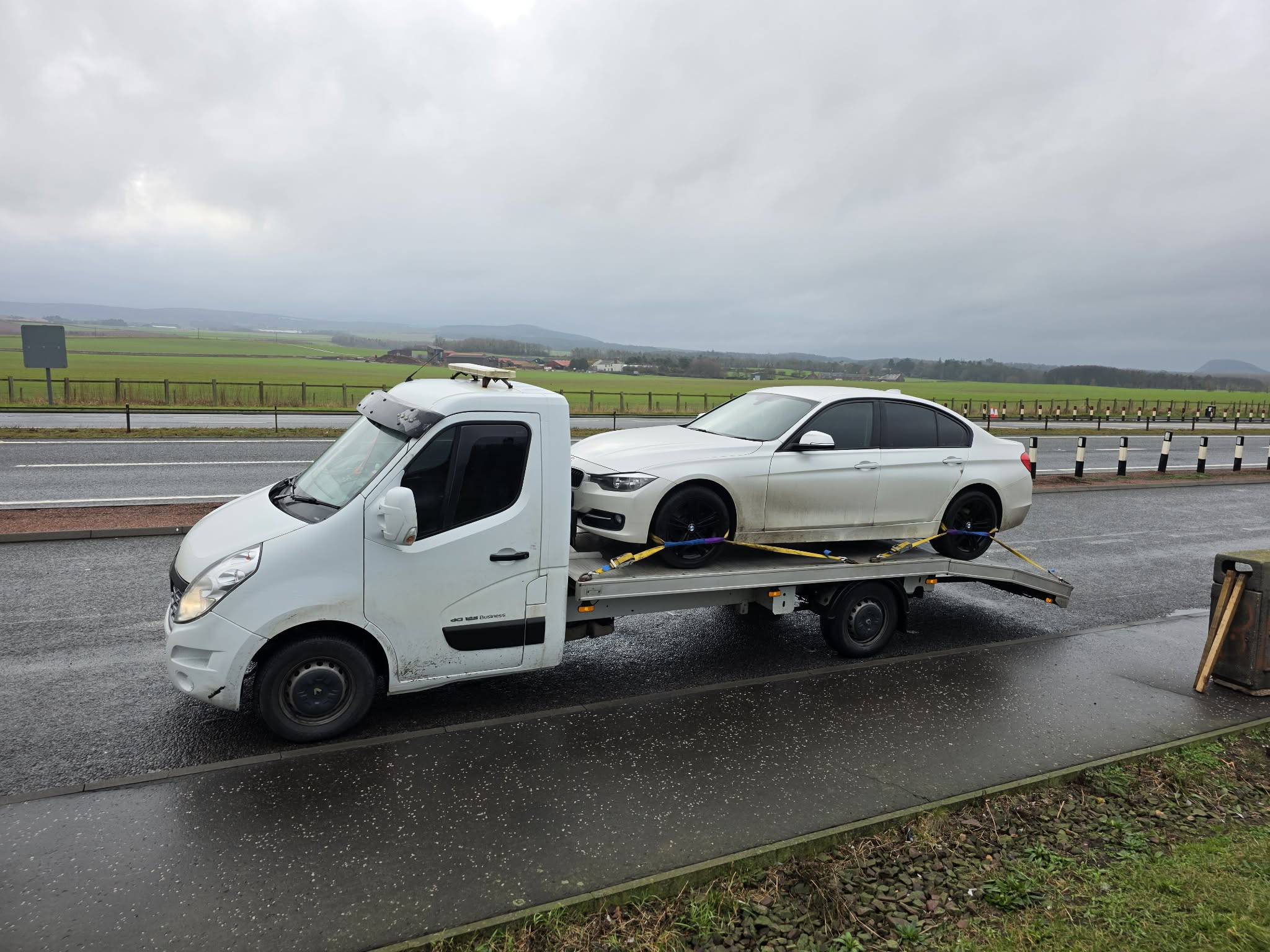 White BMW loaded on GTT flatbed on a rainy A-road with Scottish hills in the background